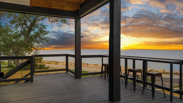 A wooden deck with a view of the sunset over the water, a fence, and two stools. - Waterfront Home w/ Views - Hot Tub + Movie Theatre (Bacliff)