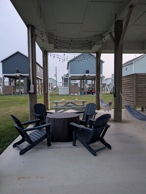 An outdoor patio area with a round table and four Adirondack chairs, surrounded by wooden beams and string lights.