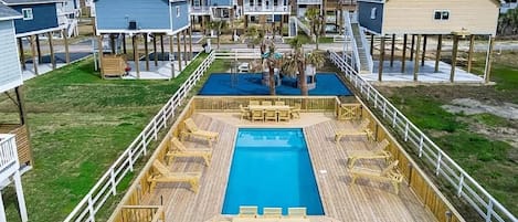 Zona de piscina amb terrassa de fusta i gandules, envoltada de cases de platja amb terrasses elevades.