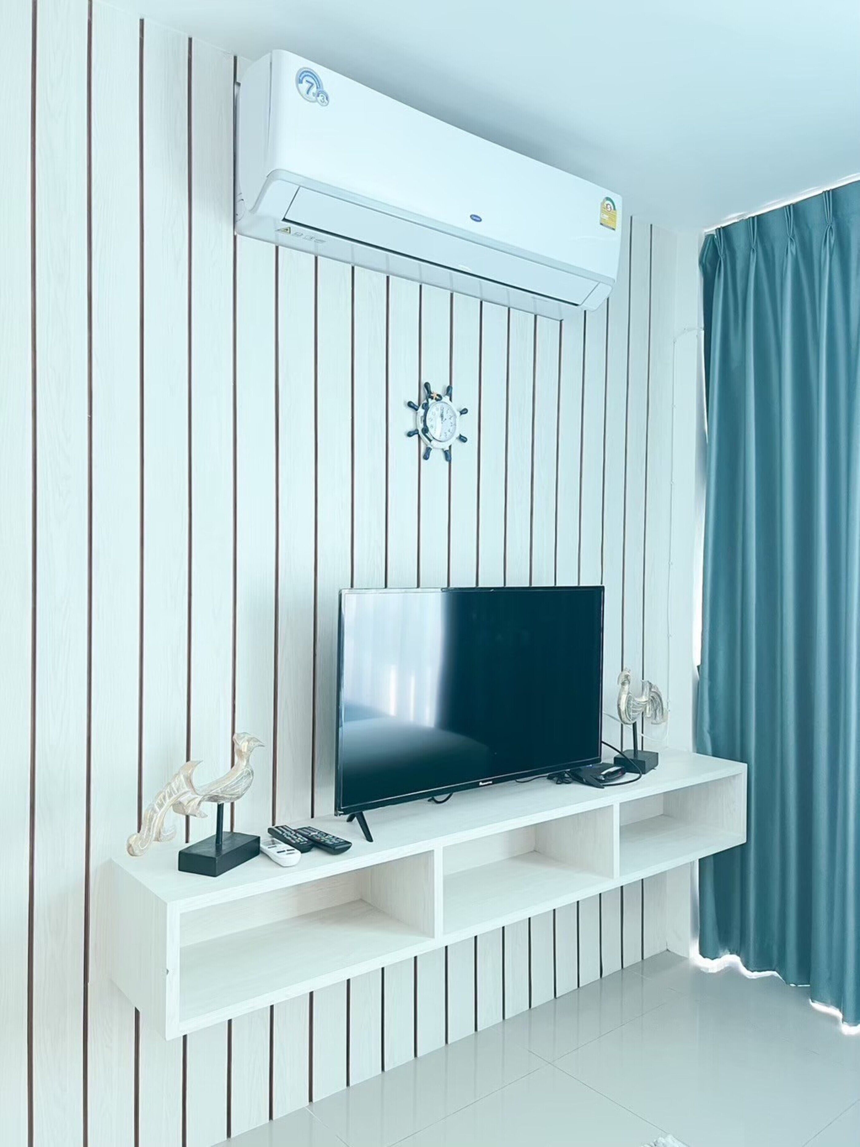 A modern living room with a flat-screen TV on a white stand, a mounted air conditioner, and a decorative wall clock.