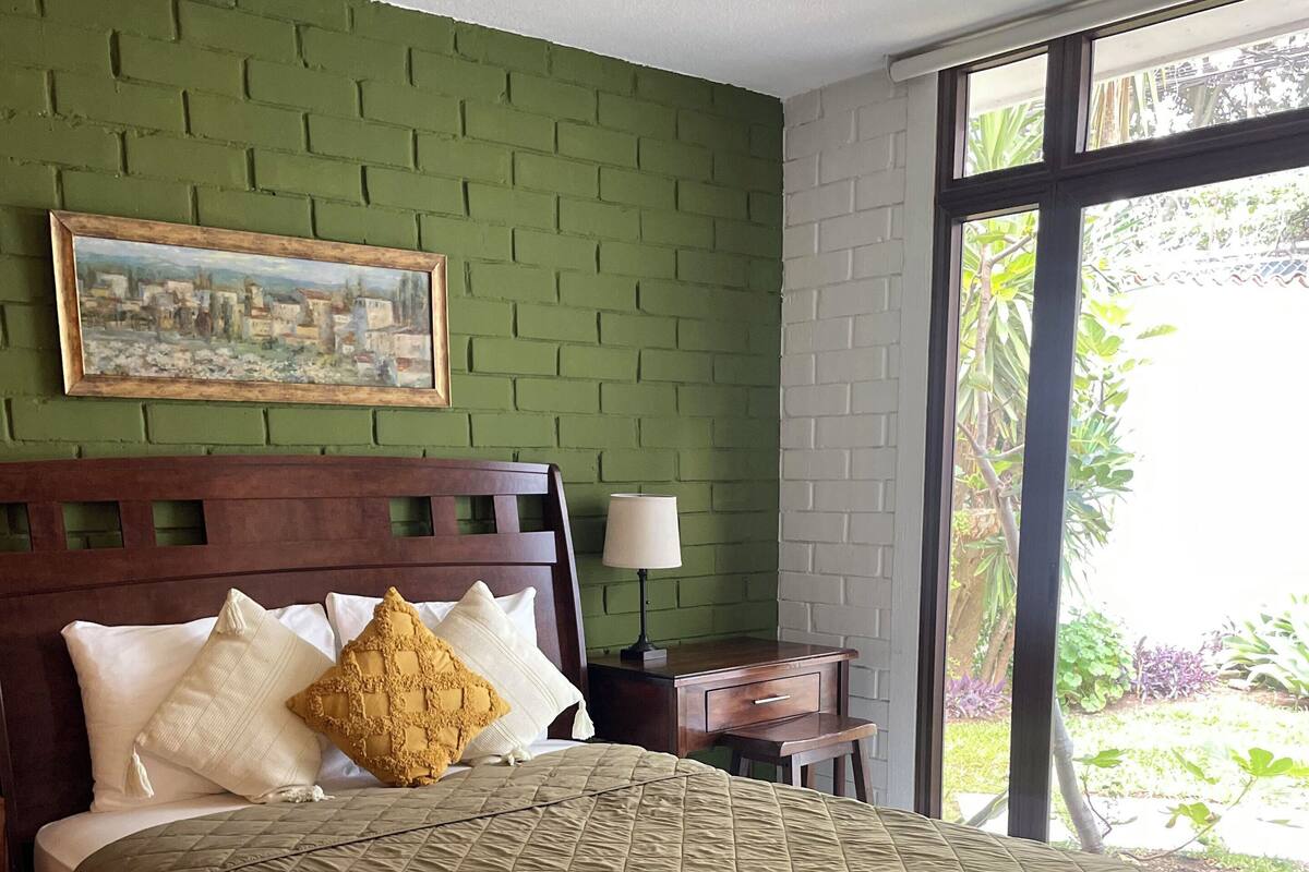 Een hotelkamer met een bed, nachtkastje, lamp en een raam met uitzicht op groen.