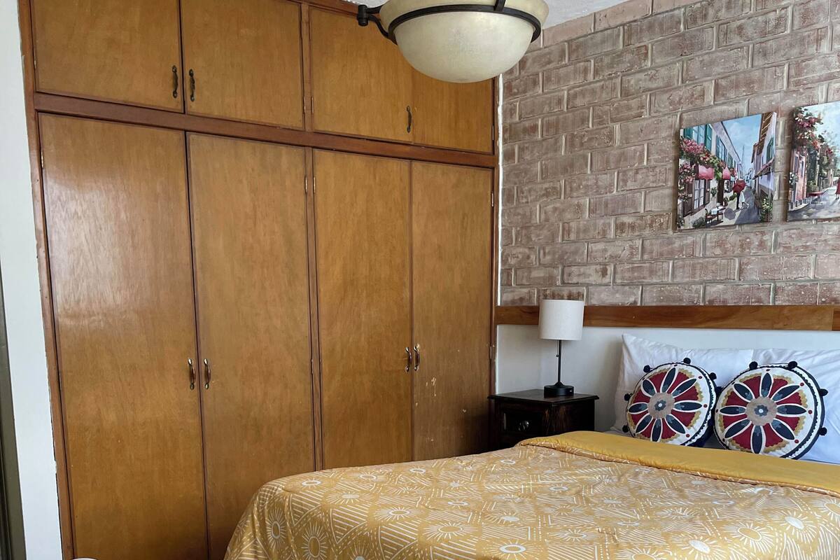 Een slaapkamer met een grote houten garderobe, een bed met een beddengoed met patroon, een bakstenen muur en een schilderij.