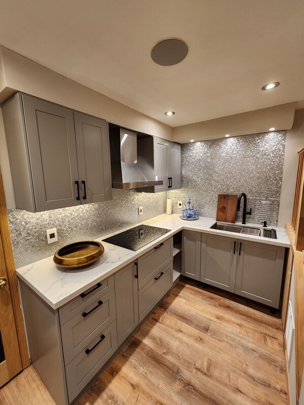 A modern kitchen with grey cabinets, a stainless steel range hood, a marble countertop, and a wooden floor.
