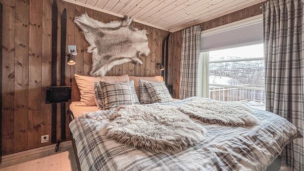 Une chambre avec un plafond en bois, un lit recouvert d’une couverture à carreaux et un tapis en fourrure.