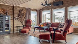 Un salon chaleureux avec un plafond en bois, une grande fenêtre avec des rideaux, un canapé en cuir et une vitrine contenant des objets décoratifs.