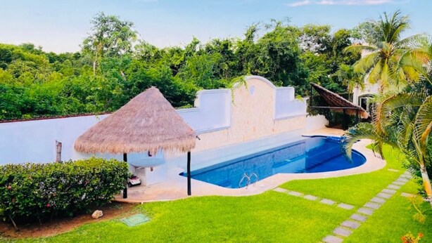 A swimming pool with a thatched roof structure, surrounded by lush greenery and a well-maintained lawn. - Basic 3 bedroom villa (Playa del Carmen)