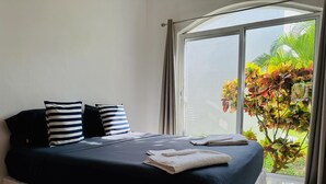 A bed with a dark blue bedspread and white pillows, a white towel on the bed, and a view of greenery through a large window. - Basic 3 bedroom villa (Playa del Carmen)