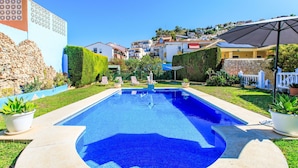 Una piscina rectangular con un diseño de mosaico azul, rodeada por un deck de concreto, plantas en macetas y una cerca blanca.
