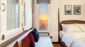 A bedroom with a bed, a bench with pillows, a window with a view of a sunset, and two framed pictures on the wall. - Cherie Bangkok Boutique Hotel (Bangkok)