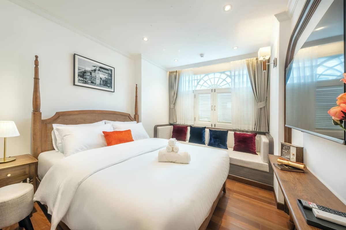 Une chambre d’hôtel avec un grand lit, une tête de lit en bois, une table de chevet avec une lampe, une fenêtre avec des rideaux et un tableau encadré au mur.