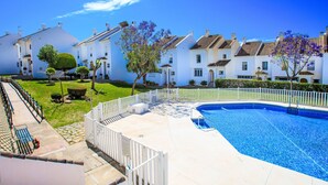 A swimming pool with a white fence, a grassy area, and a row of white buildings with brown roofs. - Los almendros by Interhome (Rincón de la Victoria)