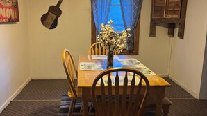 A dining area with a wooden table, chairs, a patterned rug, a guitar wall art, a window with curtains, and a hanging light fixture. - Hogan’s Hideout Bonnie Manor- Cozy Cottage to enjoy. (Eminence)
