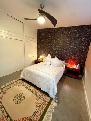 A neatly made bed with white linens, a patterned rug, a ceiling fan, and a wall with a dark, abstract design.