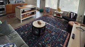 A modern living room with a large rug, a sofa, a coffee table, and a kitchen area with wooden cabinets and a stove.