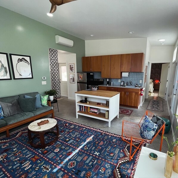 A modern living room with a sofa, coffee table, and patterned rug. A kitchen with wooden cabinets and a dining area is visible in the background.