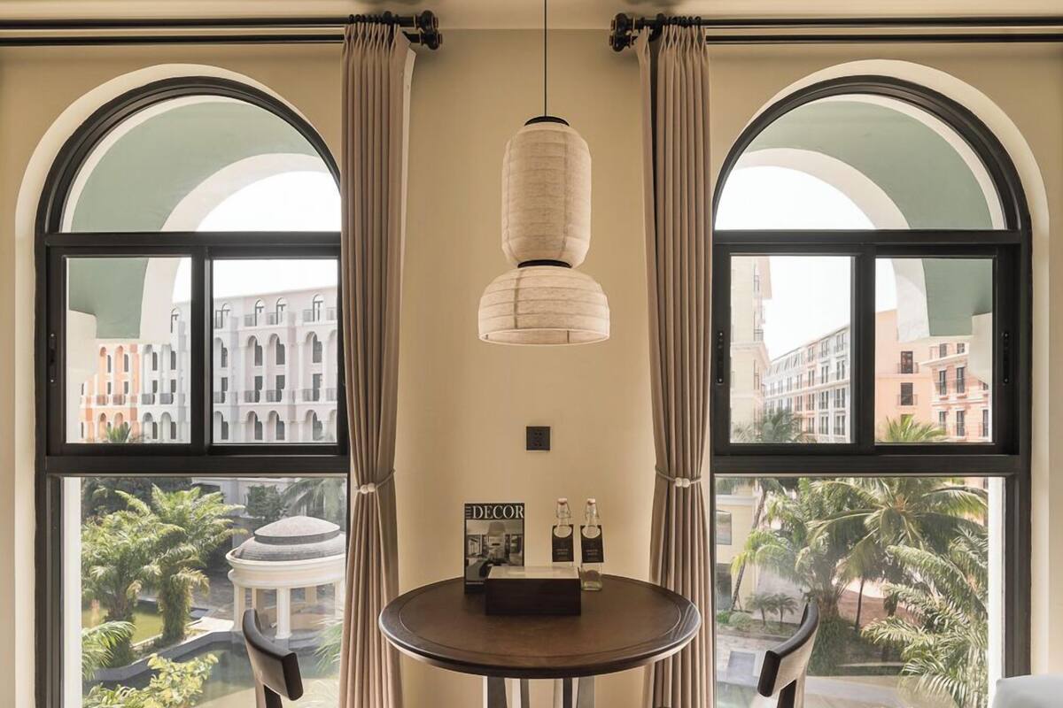 Un comedor con una mesa redonda, dos sillas, una lámpara colgante y ventanas grandes con cortinas.