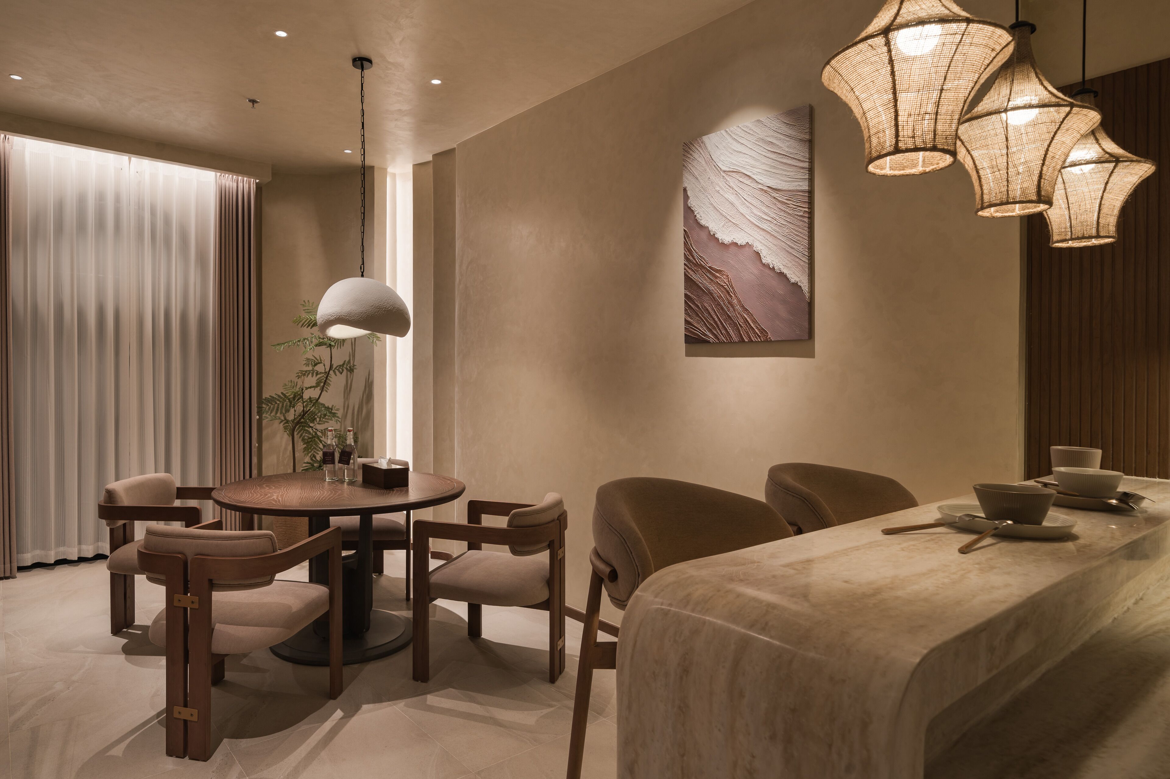 A modern hotel room with a round dining table, four chairs, a large bed, and a hanging light fixture.
