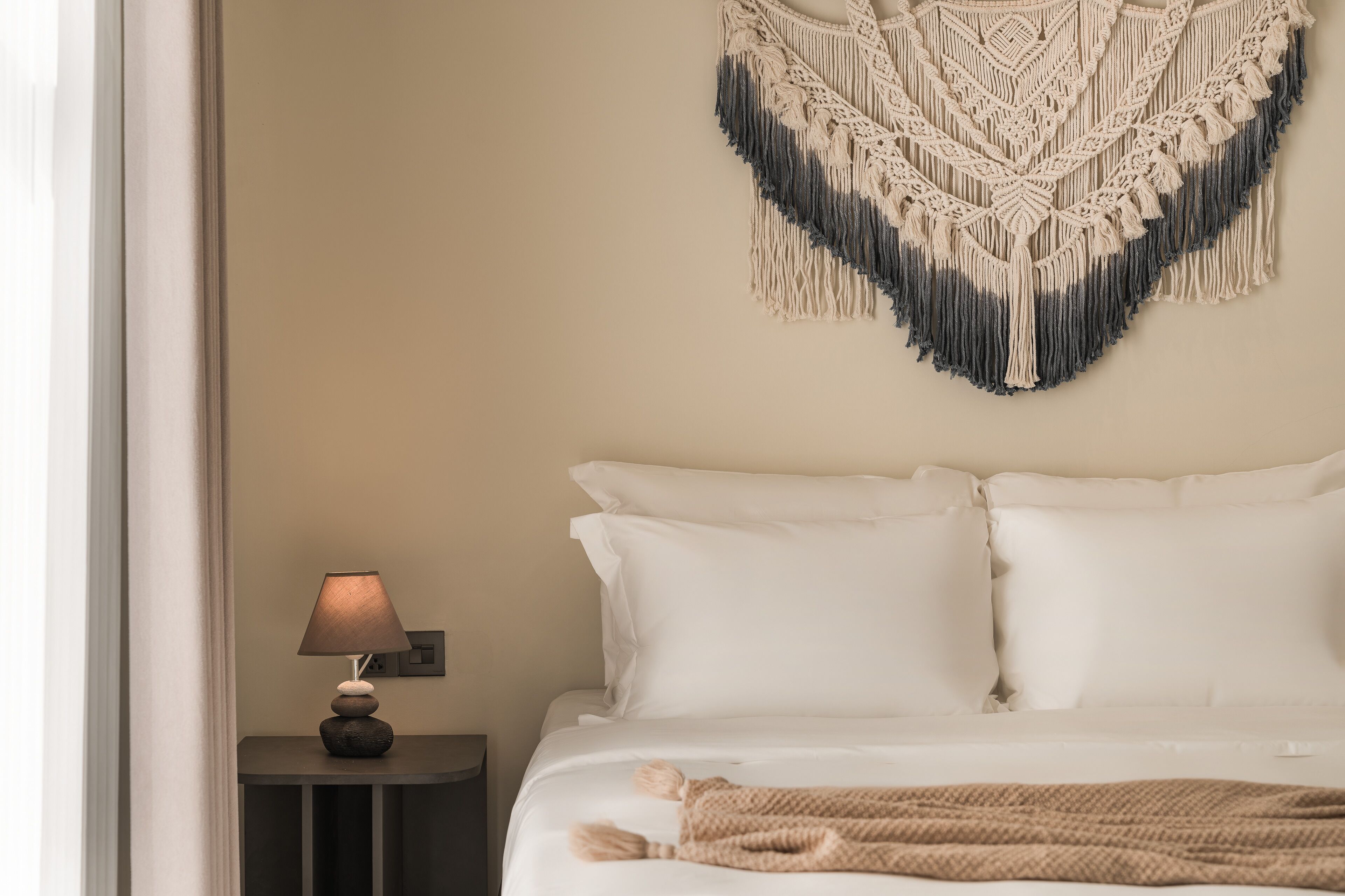 A neatly made bed with white linens, a beige macrame wall hanging, and a small table with a lamp beside it.