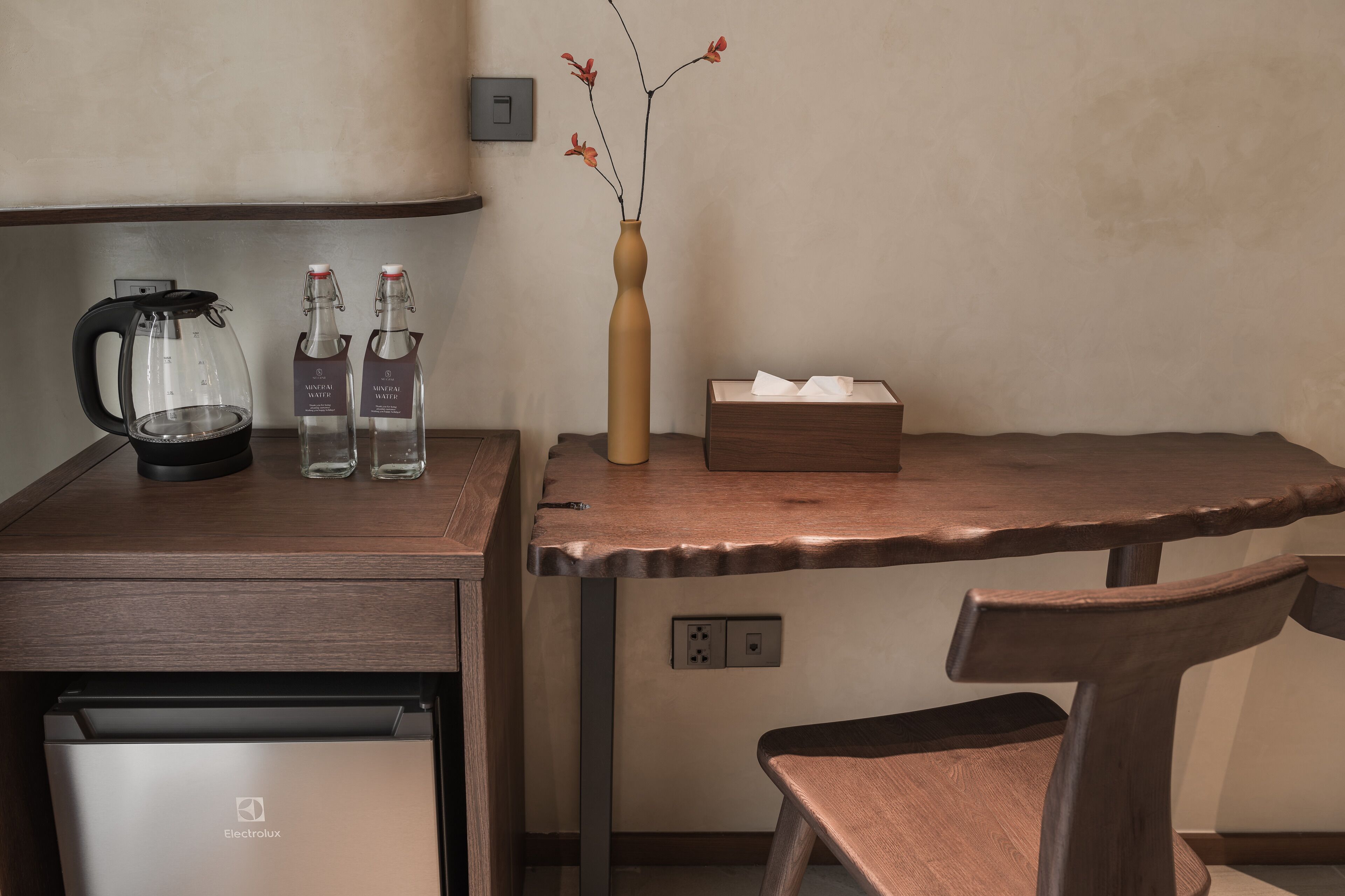 Una kitchenette con cafetera, dos botellas de agua, un florero con flores, una caja de pañuelos y una silla de madera.