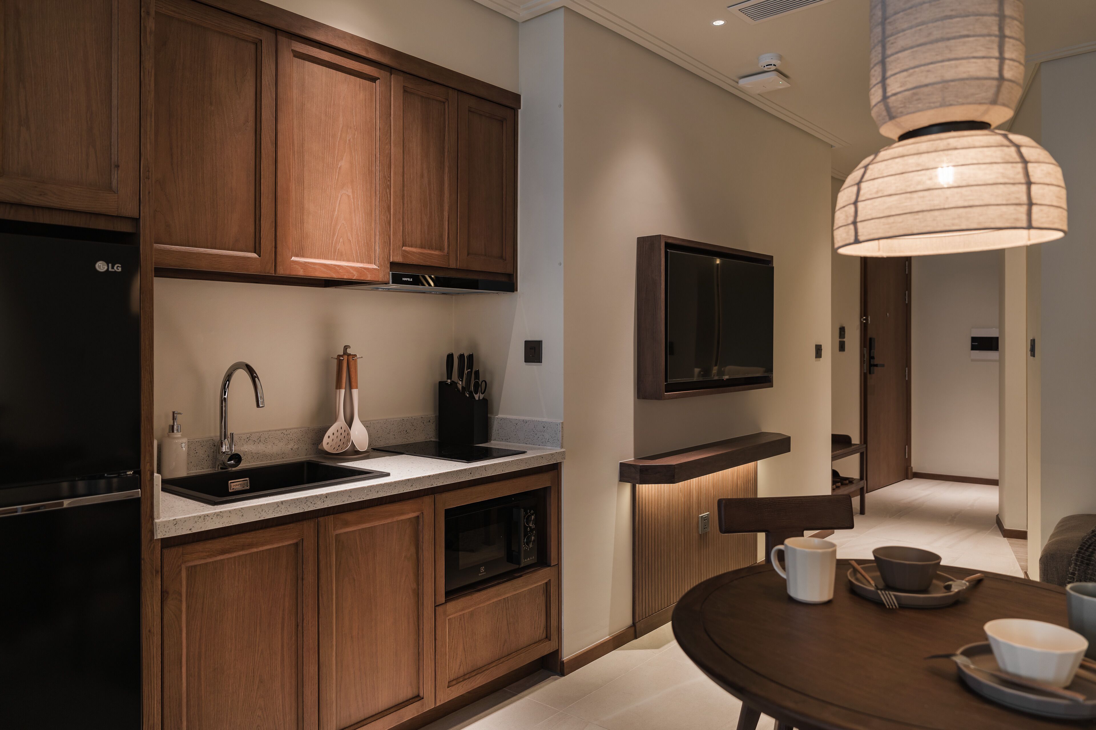 A modern kitchen with wooden cabinets, a built-in dishwasher, a stainless steel sink, and a flat-screen TV mounted above the counter.
