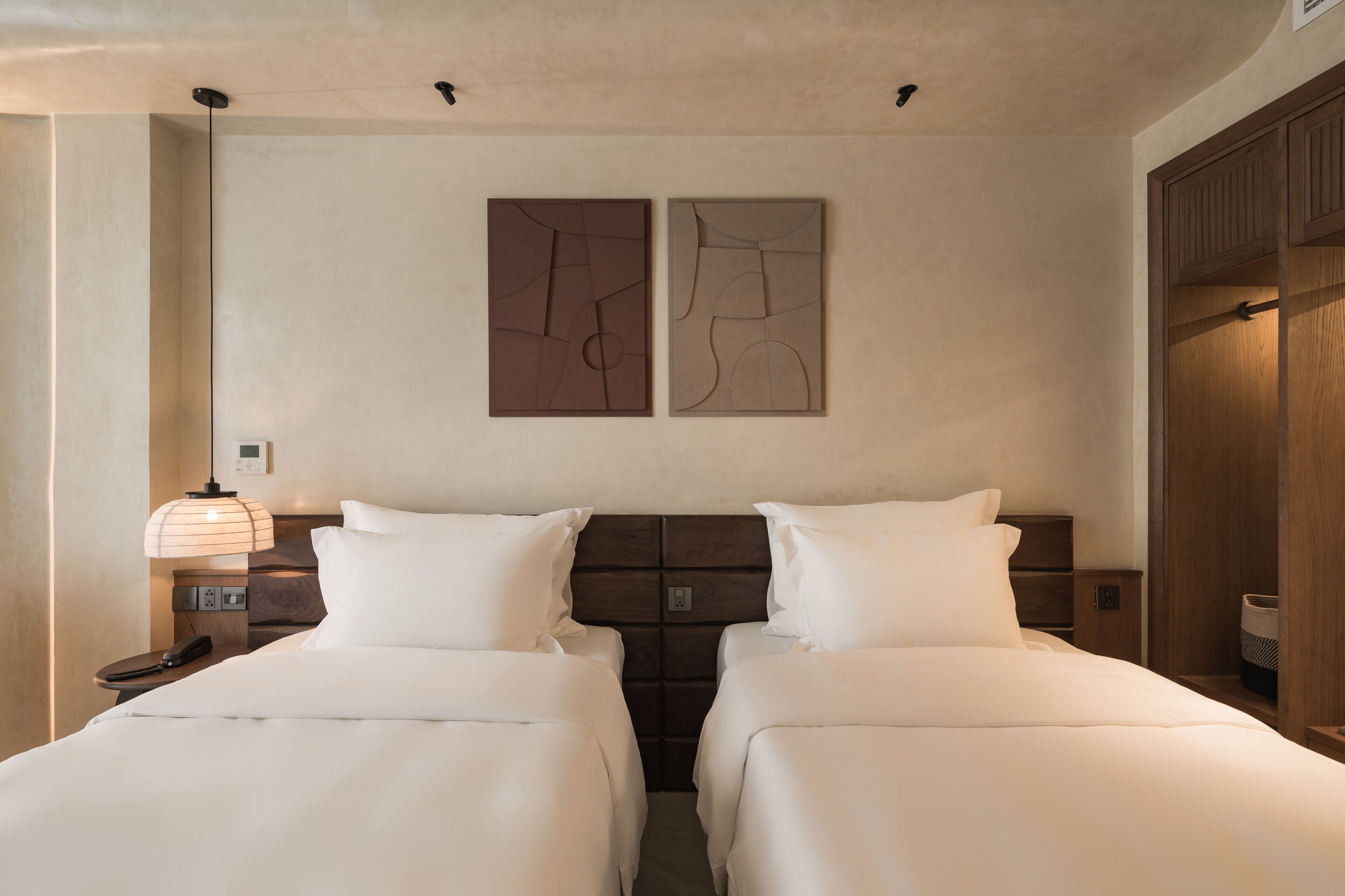Una habitación de hotel con dos camas, una mesita de noche con lámpara y un armario de madera.