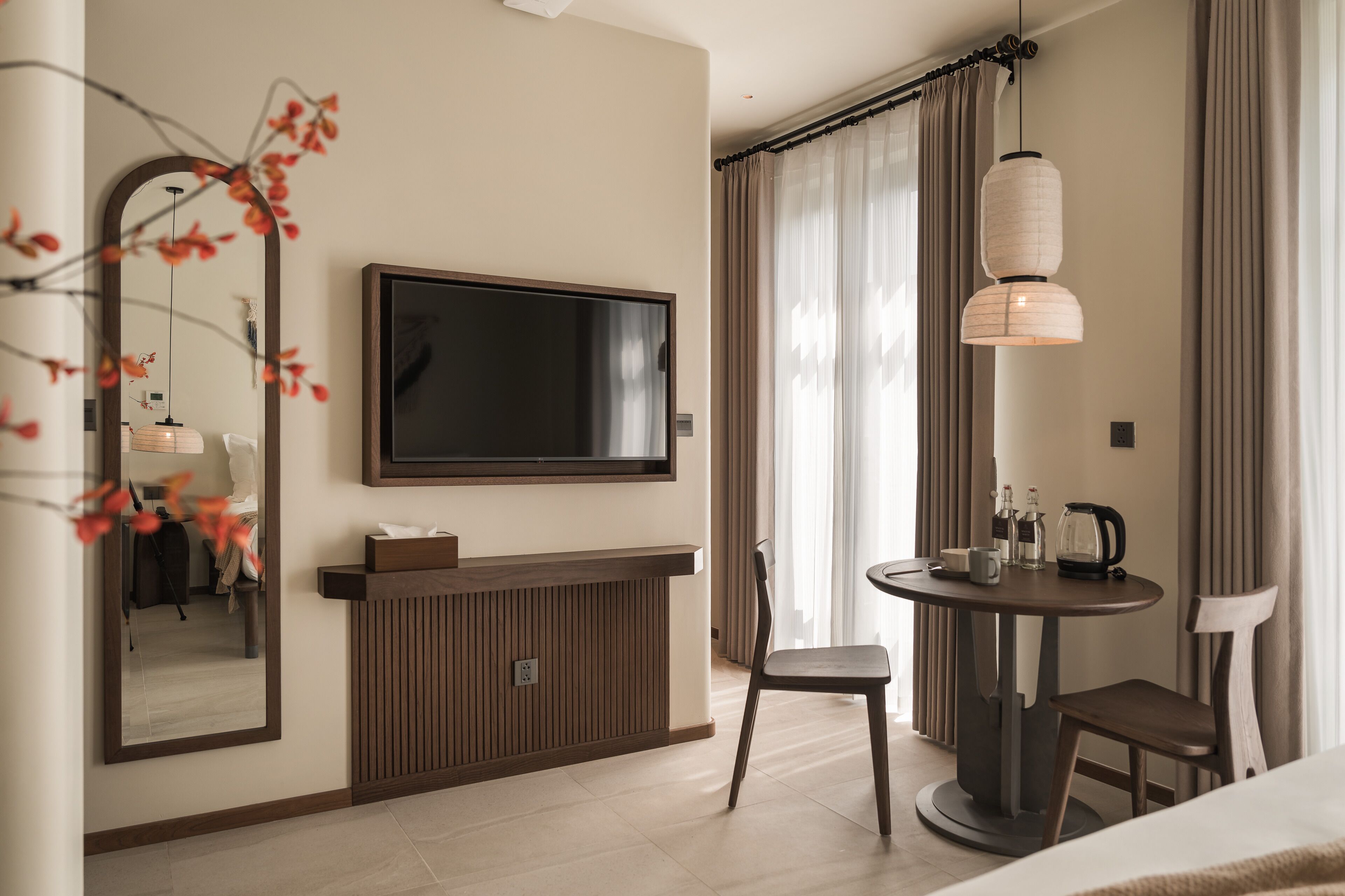 A modern hotel room with a flat-screen TV, a wooden console table, a round dining table with chairs, and a large mirror.