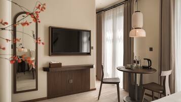 A modern hotel room with a flat-screen TV, a wooden console table, a round dining table with chairs, and a large mirror.