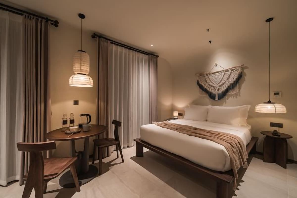 A modern hotel room with a large bed, a small round table with chairs, a bedside table with a lamp, and a decorative wall hanging.