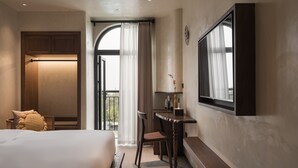 Habitación de hotel con cama, escritorio, silla, televisor y balcón con cortinas.
