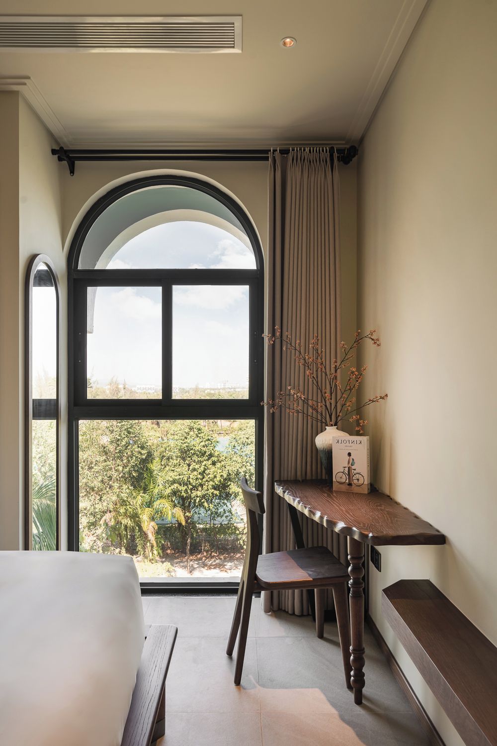 A bedroom with a large arched window, a bedside table with a vase, a chair, and a bench.