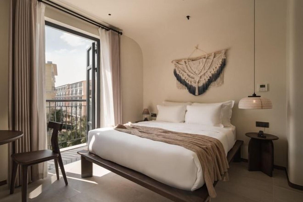 A hotel room with a large bed, a bedside table, a chair, a hanging macrame wall decoration, and a view through a large window.
