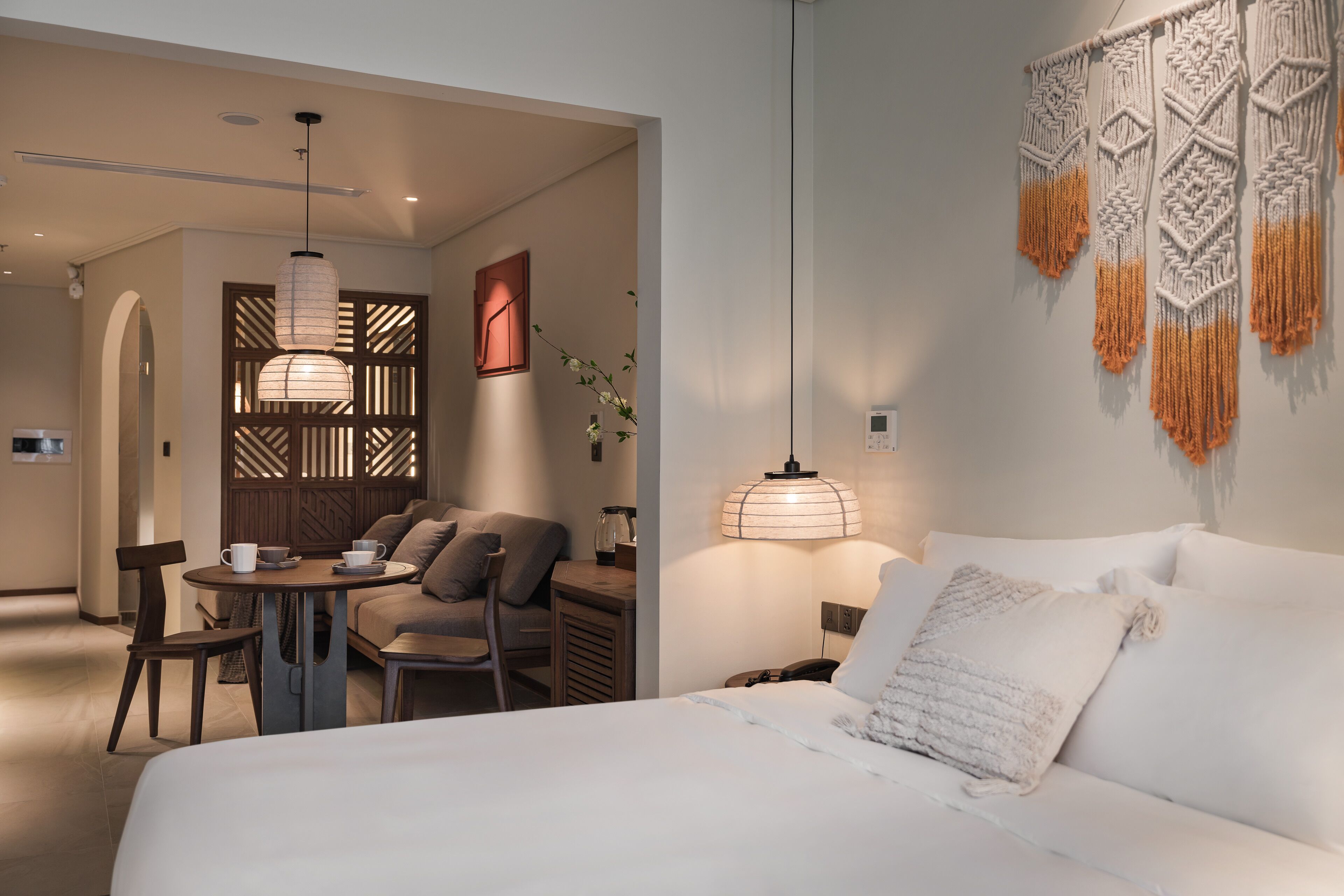 A modern hotel room with a bed, a seating area with a table and chairs, a decorative wall hanging, and a unique lighting fixture.