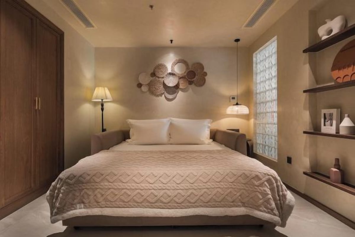A bedroom with a large bed, two bedside lamps, a wall-mounted decorative piece, and built-in shelving with decorative items.