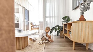 Lapsi leikkii puisilla palikoilla valoisassa olohuoneessa, jossa on suuri ikkuna, pyöreä puupöytä ja puinen kaappi hyllyineen.