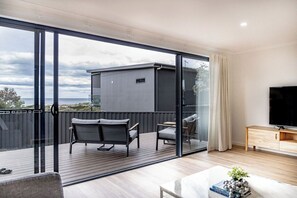 Un salon moderne doté d’une grande porte-fenêtre coulissante donnant sur un balcon. La pièce est équipée d’un téléviseur à écran plat, d’une table basse en bois et d’un canapé.