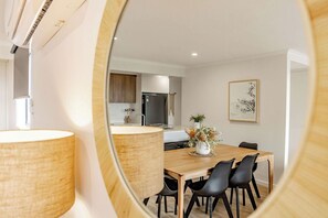 Un espace repas moderne avec une table en bois et des chaises noires, une cuisine en arrière-plan et une œuvre d’art encadrée au mur.