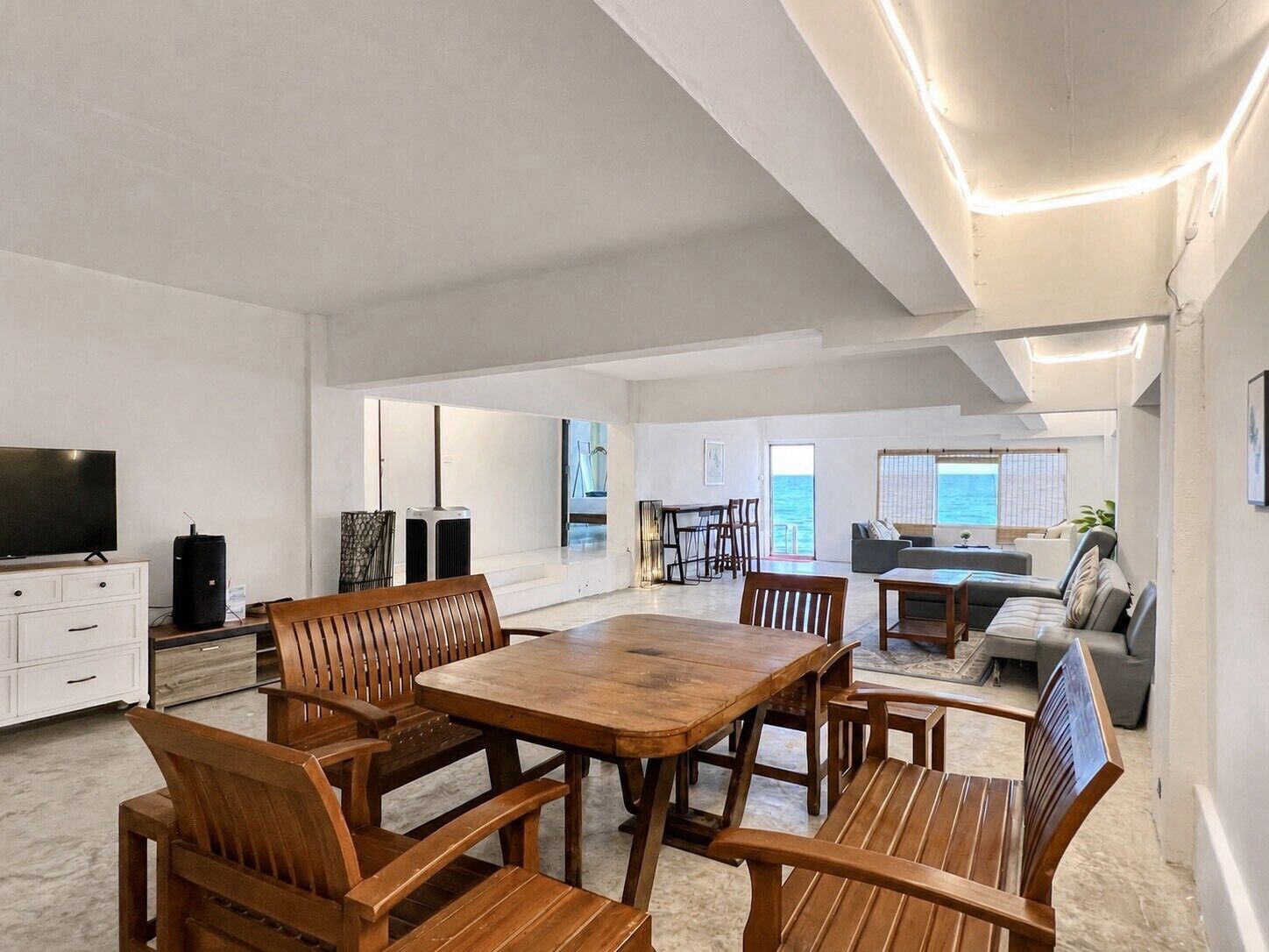 A spacious living area with a wooden dining table and chairs, a flat-screen TV, and a view of the sea through large windows.