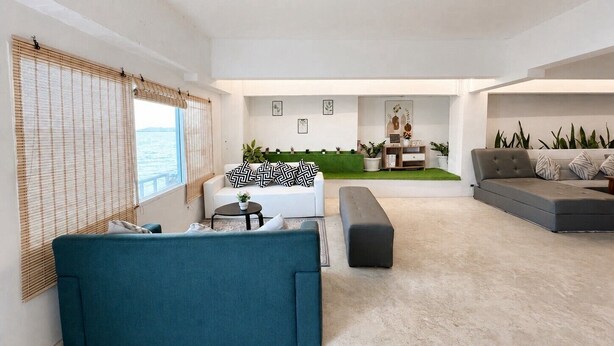 Una sala de estar moderna con un sofá, una mesa de centro y un sofá seccional. Hay una mesita con una planta y un marco de fotos. La habitación tiene un ventanal grande con persianas.