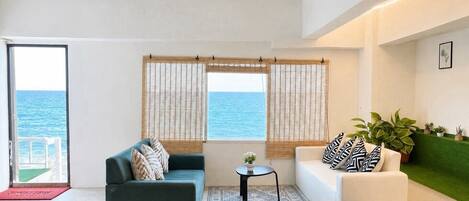 A modern living room with a sofa, ottoman, and a small table, a view of the sea through large windows, and a green wall with plants.