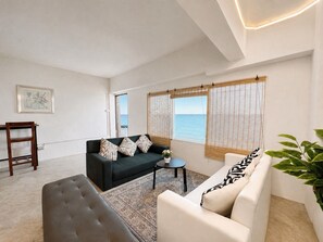 Un salon moderne avec un canapé, un fauteuil et une petite table. Une fenêtre offre une vue sur la mer.