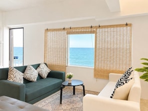 A room with a green sofa, a white armchair, a small table with a plant, and a view of the sea through bamboo blinds. - GAO Check In Phala Beachfront (Ban Chang)
