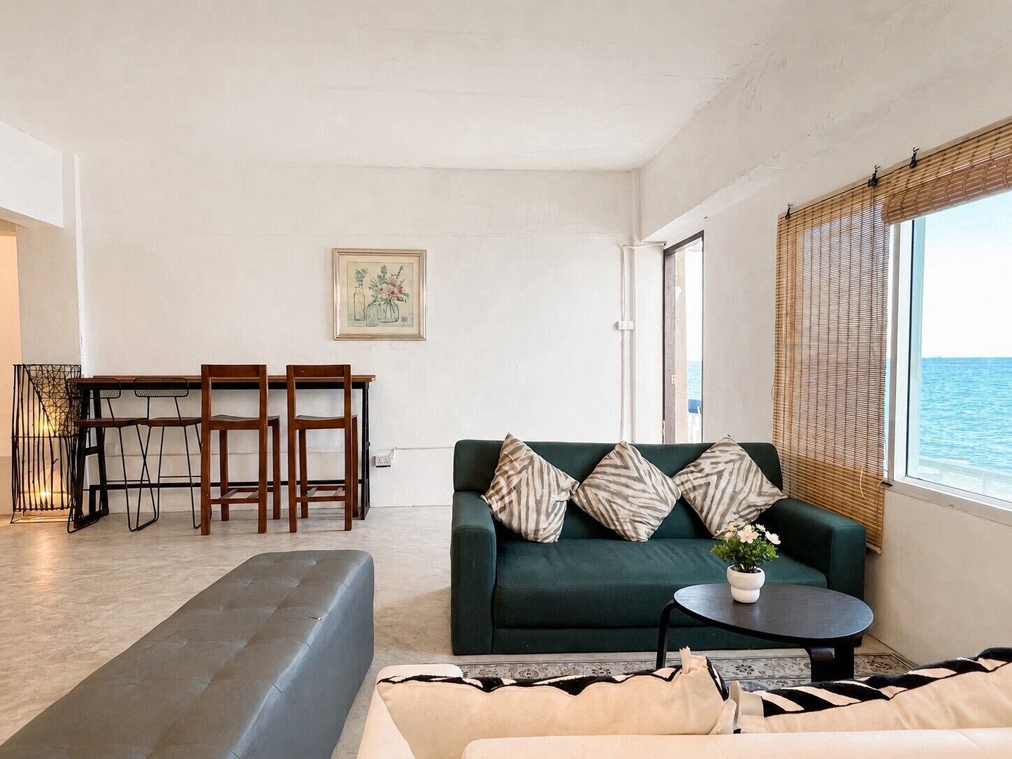A living room with a sofa, a dining table with chairs, and a view of the sea through a window.