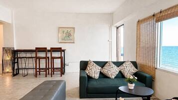 A living room with a sofa, a dining table with chairs, and a view of the sea through a window.