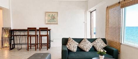 A living room with a sofa, a dining table with chairs, and a view of the sea through a window.