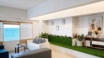 A modern living room with a sofa, ottoman, and a small table with a plant. There is a green wall with a shelf and a sign, and a window with a view of the sea.