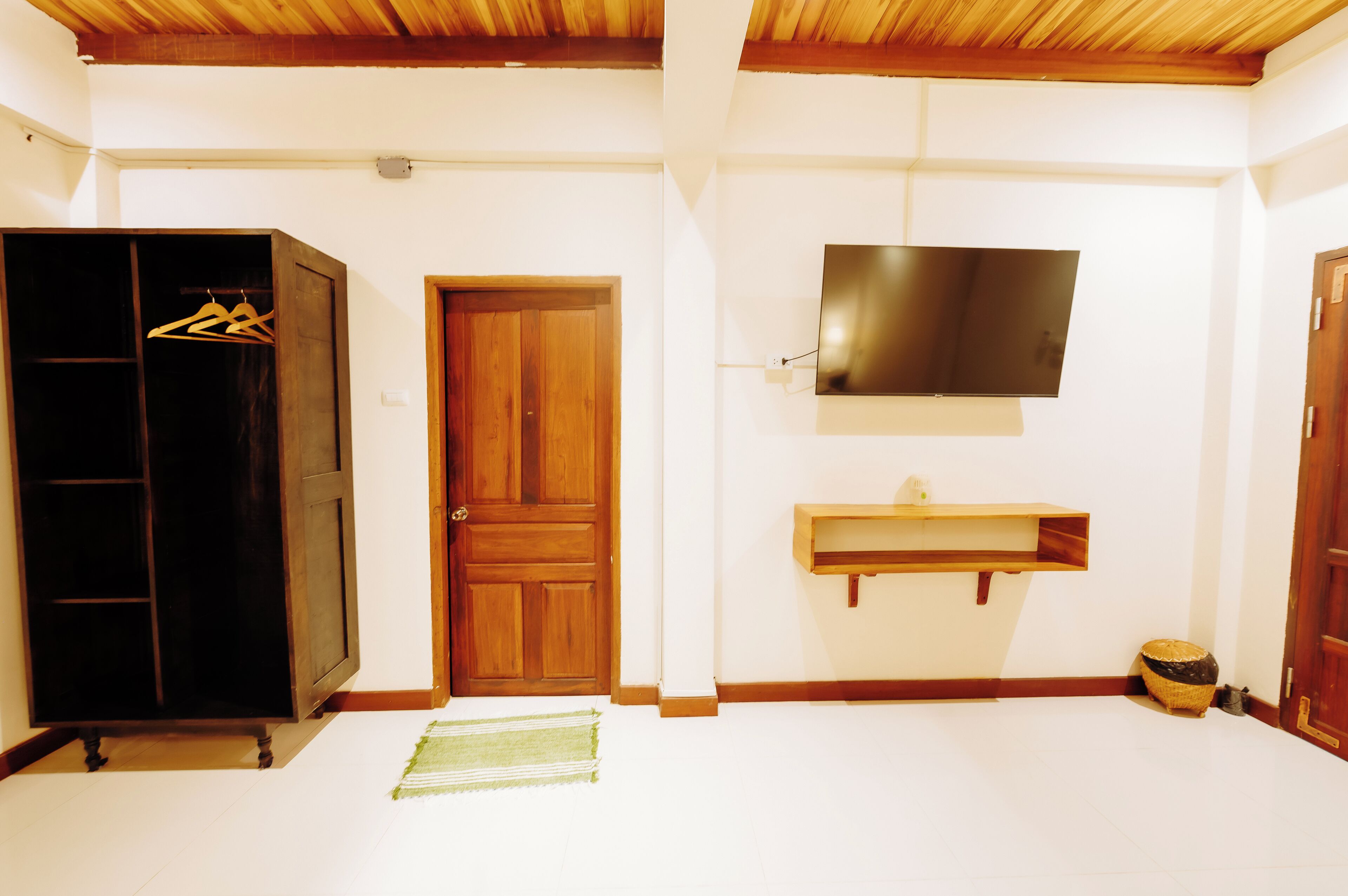 A room with a wooden wardrobe, a door, a television mounted on the wall, and a small shelf.