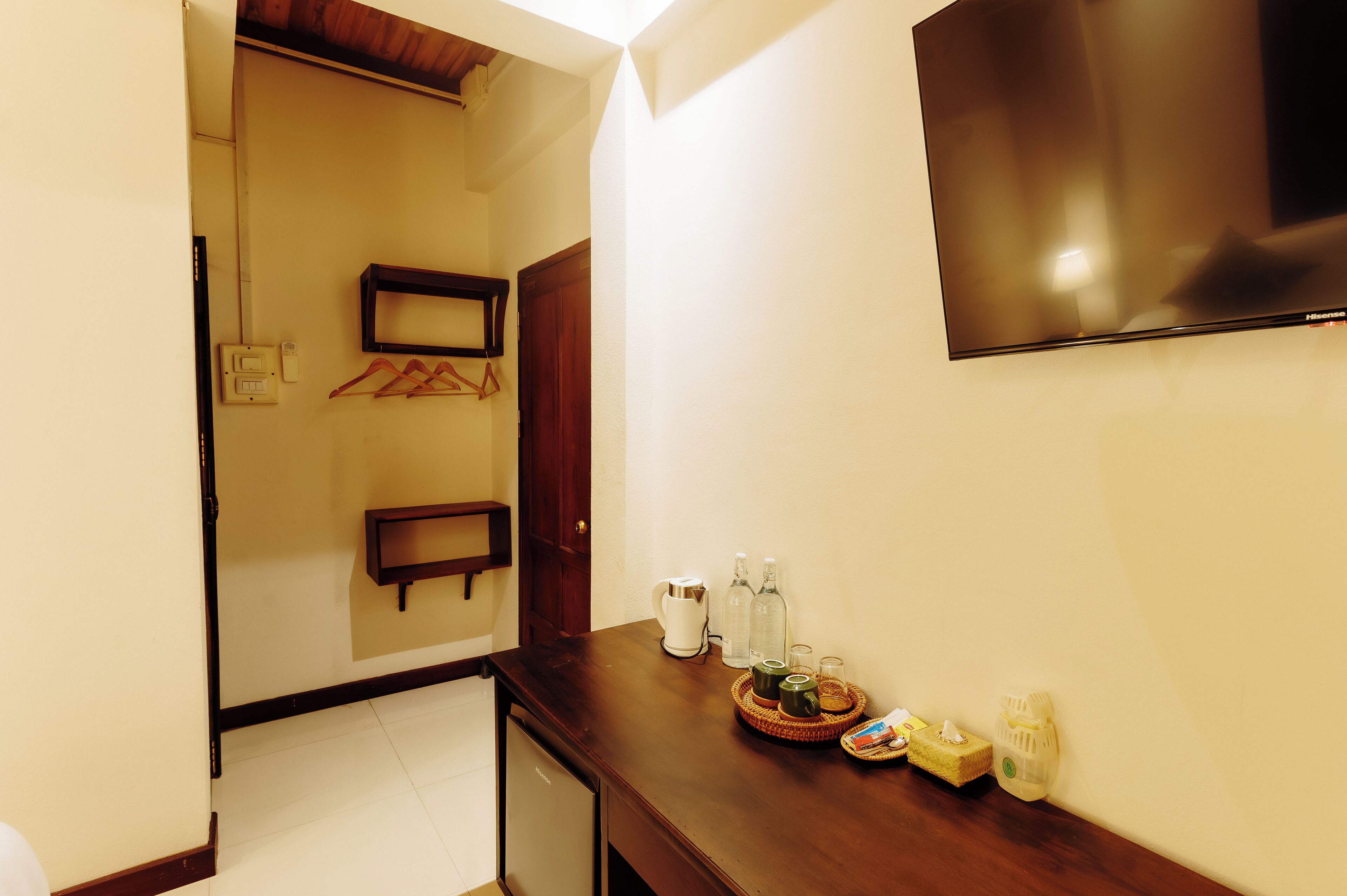 A hotel room with a wooden desk, a flat-screen TV, a small table with bottles and glasses, and a wall-mounted shelf with hangers.