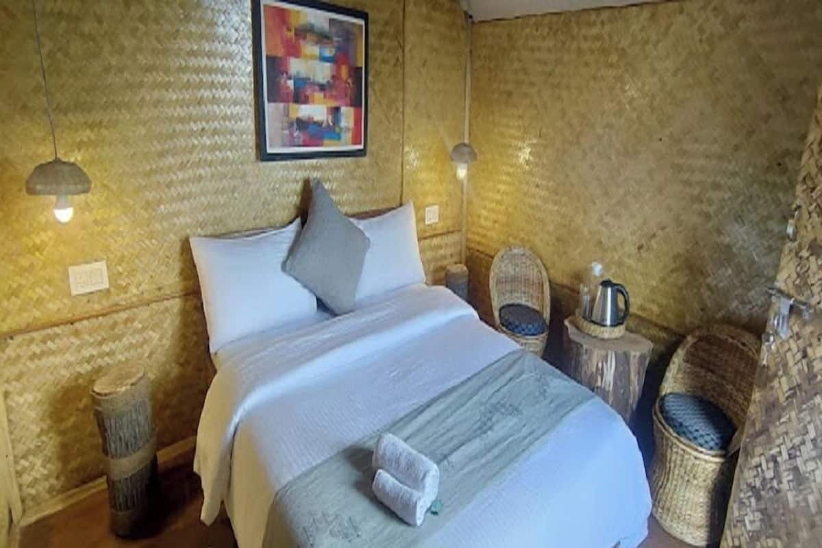 A bed with a white comforter and pillows, a small bedside table with a lamp, a woven basket, and a framed abstract painting on the wall.