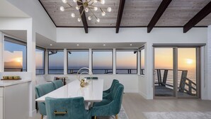Un comedor moderno con una mesa blanca y sillas color turquesa, ventanas amplias con vistas a la puesta de sol y techo de madera.