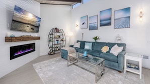 A modern living room with a large flat-screen TV, a fireplace, a teal sofa, and a glass coffee table. - Oceanfront/ Stunning Views/ Hot Tub/ Contemporary (Manchester)